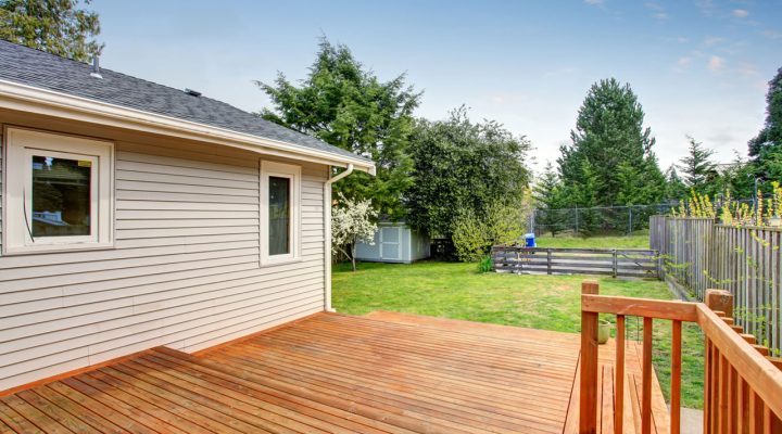 A large, clean wooden deck with a natural finish extends from the side of a beige-sided home, offering an open view of a lush green backyard with trees, a shed, and a fenced area. The deck features simple railings and provides ample space for outdoor living.