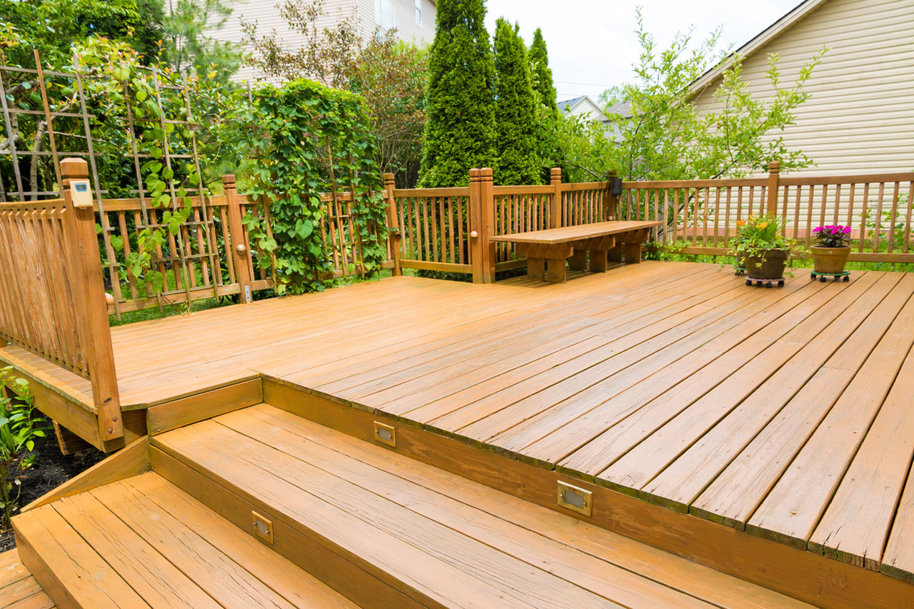 A beautifully crafted multi-level wooden deck featuring a built-in bench, decorative railing, and integrated step lighting. The deck is surrounded by lush greenery, including ivy-covered trellises and tall evergreens, creating a private and inviting outdoor space.