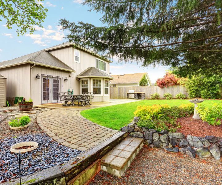 A well-designed backyard with a winding paver path leading through a mix of gravel, mulch, and rock gardens. The space includes a birdbath, flower beds, a patio area, and mature trees, offering a harmonious blend of hardscaping and natural beauty.