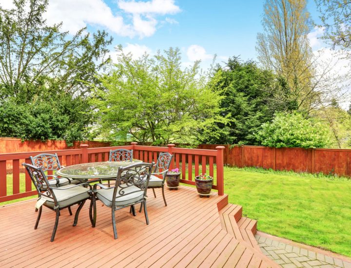A spacious wooden deck with rich-toned planks and red railings, furnished with a glass-top table and metal chairs. The deck overlooks a well-maintained lawn and is enclosed by a matching wood fence, offering a perfect space for outdoor dining and relaxation.