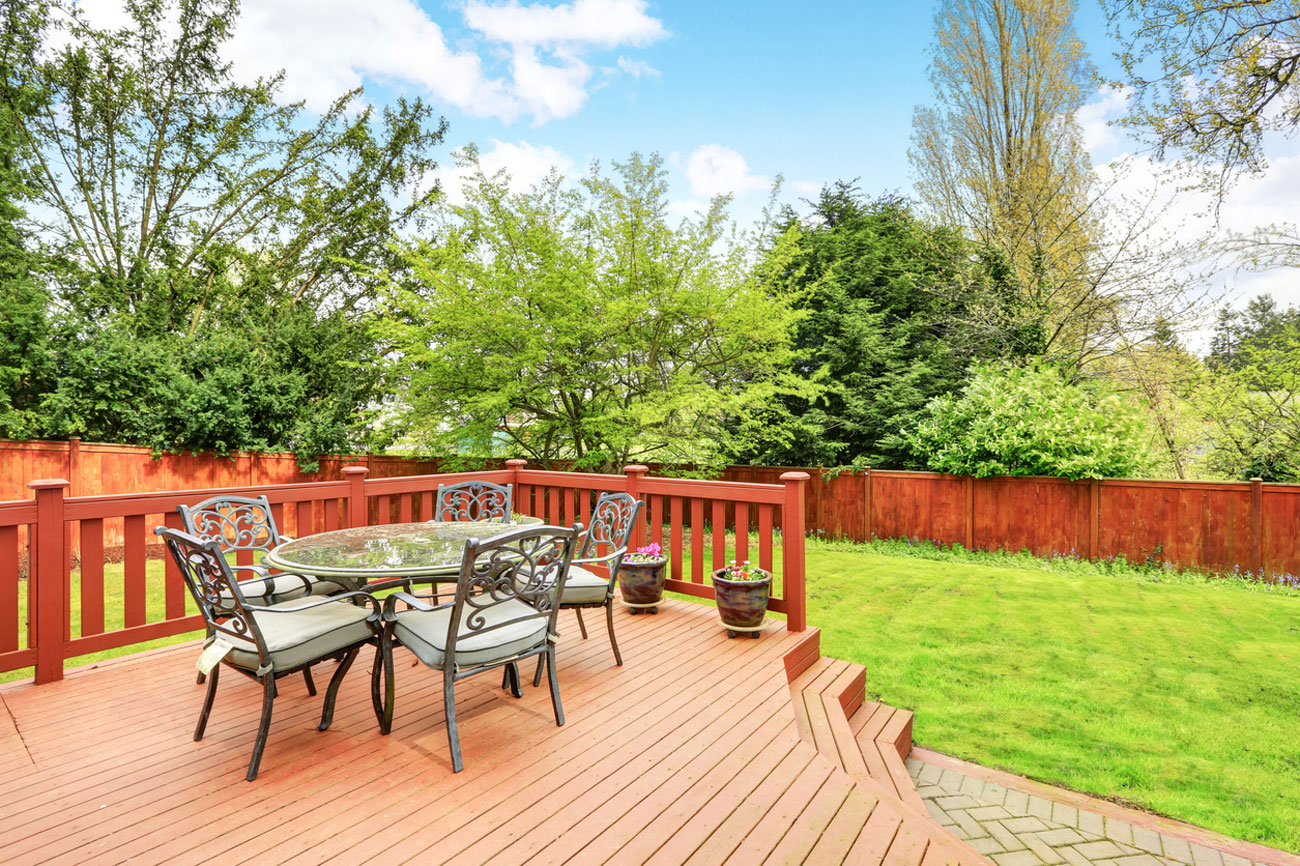 A spacious wooden deck with rich-toned planks and red railings, furnished with a glass-top table and metal chairs. The deck overlooks a well-maintained lawn and is enclosed by a matching wood fence, offering a perfect space for outdoor dining and relaxation.