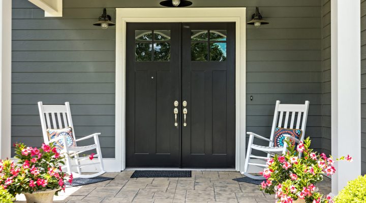 A welcoming front porch with a stone paver floor, flanked by two white rocking chairs and vibrant pink flowering plants in urns. The porch features a black double door with glass panels, framed by gray siding and white trim, creating a classic and cozy entrance.