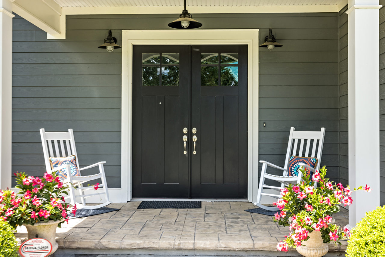 A covered concrete patio supported by white posts and a wooden roof, featuring a hammock in the center. The space is surrounded by a well-maintained landscape with colorful shrubs, trees, and a distant red play structure, ideal for relaxation and family gatherings.