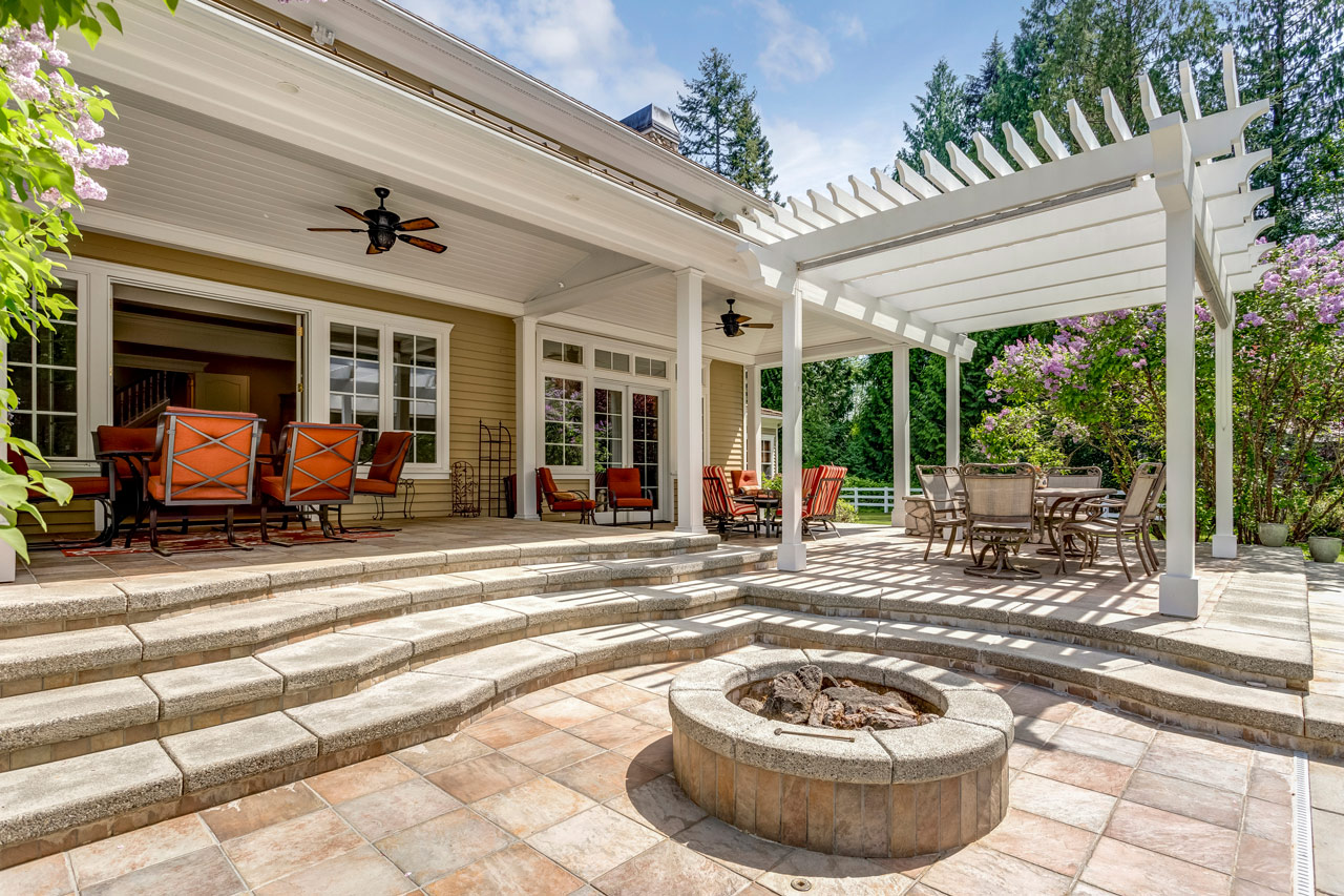 A spacious backyard patio with a white wooden pergola providing shade over a seating area, centered around a round stone fire pit. The space features tiered stone steps, tile flooring, and comfortable outdoor furniture, surrounded by blooming lilac trees and lush greenery.