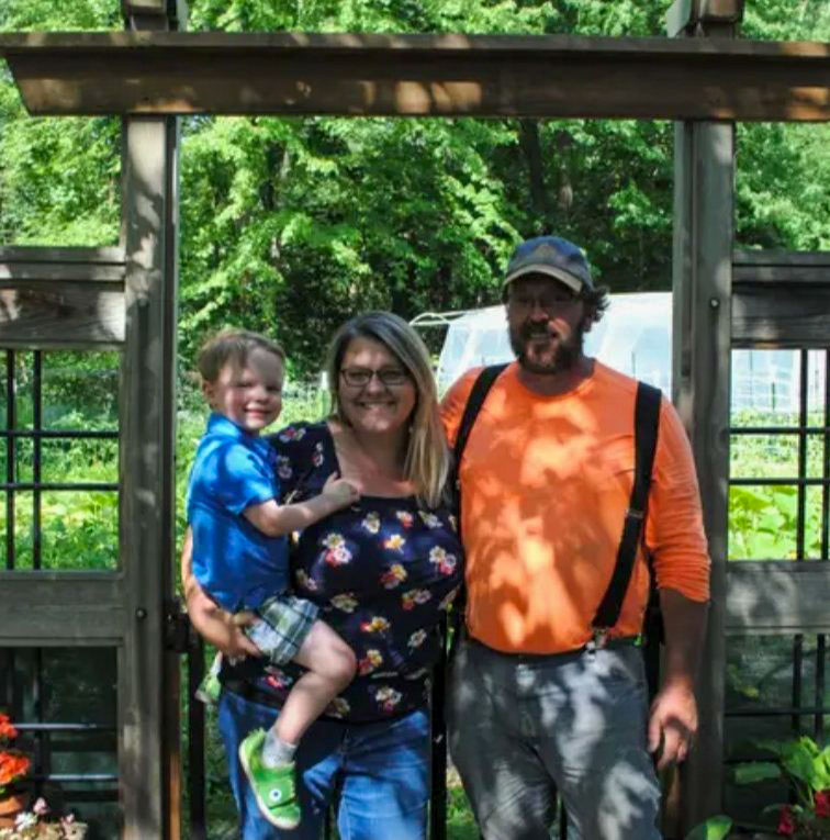 A smiling family of three—mother, father, and young child—standing together under a wooden pergola in a vibrant garden. The background features green foliage and a greenhouse, capturing a moment of pride and connection in their outdoor space.